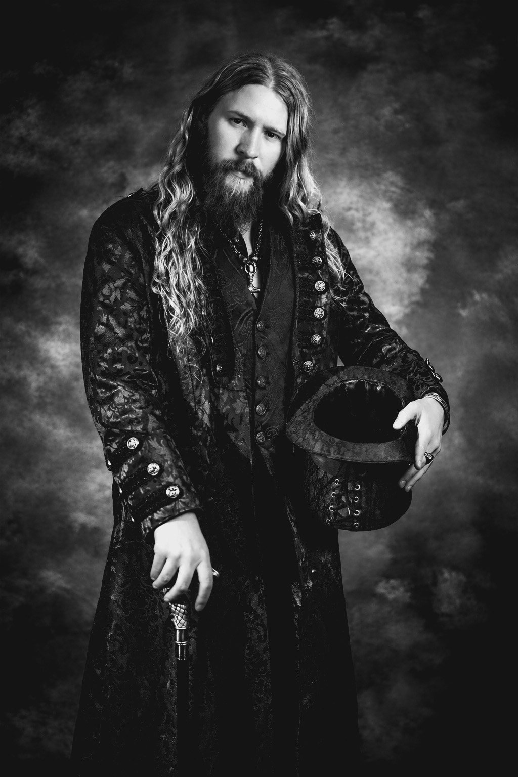 Black-and-white studio portrait of a long-haired, bearded man in a dark brocade coat and waistcoat, holding a top hat and a silver-handled cane