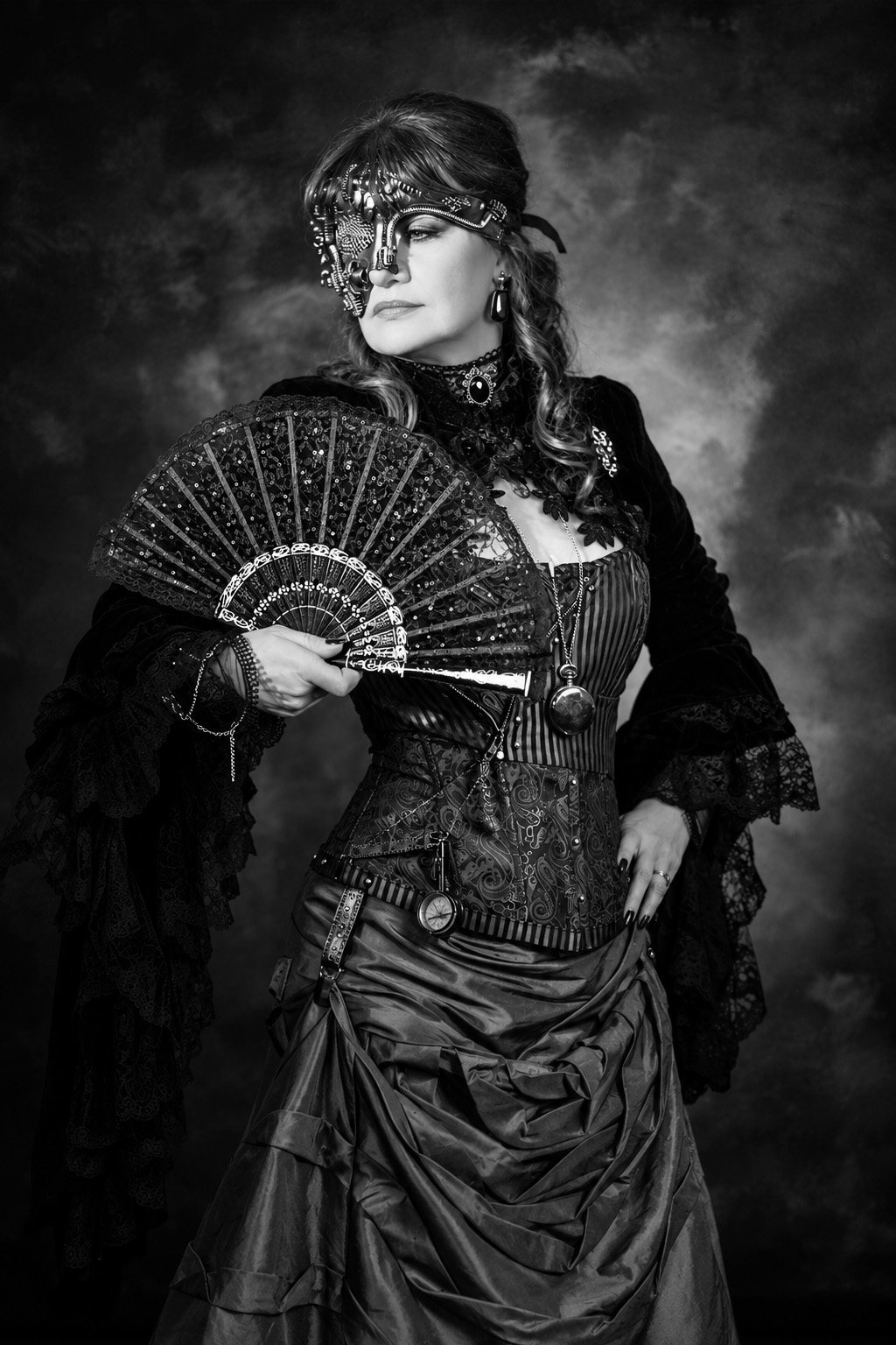 Black-and-white portrait of a woman wearing a silver mechanical half-mask and holding a black lace fan, in a corset with a pocket watch on a chain