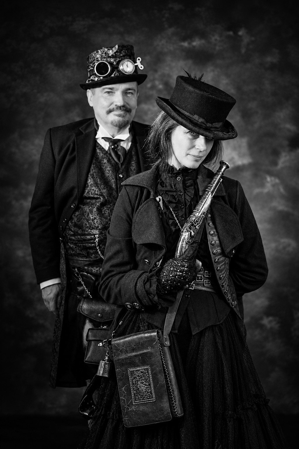 Black-and-white portrait of a man in a goggled top hat standing behind a woman in a black top hat who holds an ornate prop pistol