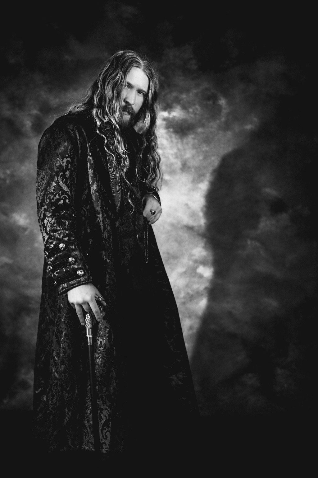 Black-and-white portrait of a long-haired, bearded man in a long brocade coat resting a hand on a cane, against a dark stormy backdrop