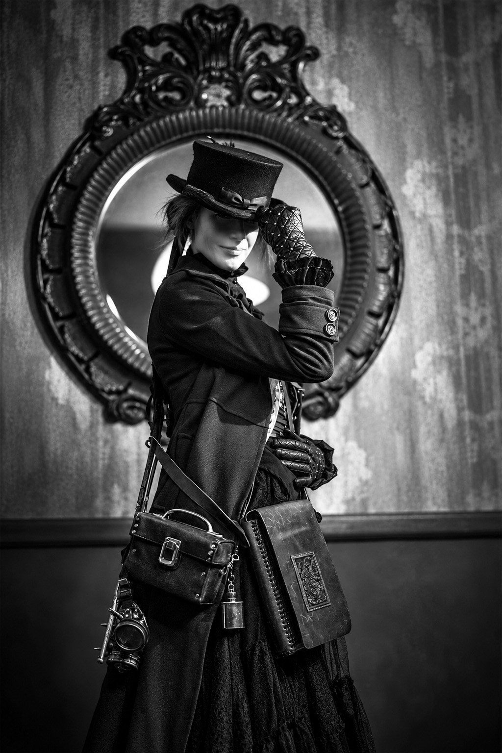 Black-and-white portrait of a person in a top hat and long dark coat, tipping the hat in front of a large ornate round mirror, with leather bags and goggles at their side
